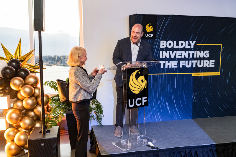 Man in a suit at a clear podium presents a glass award to a woman on a stage with a UCF backdrop and gold balloon arch outside the window.