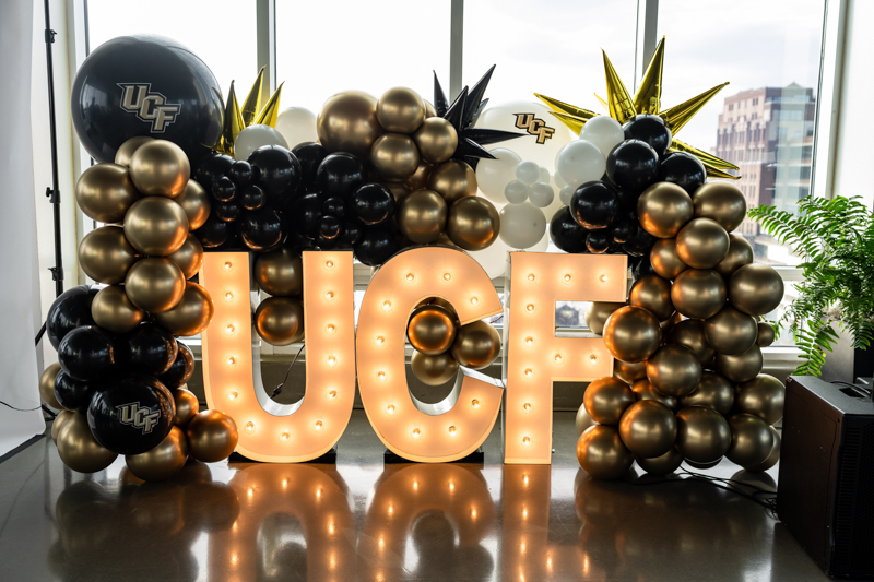 Large illuminated 'UCF' letters surrounded by black, gold, and white balloons in a bright room for a celebration. Includes UCF logos on some balloons.