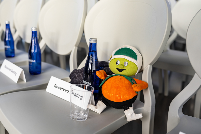 Colorful plush toy with a green hat sits on a gray conference chair beside blue bottles and a 'Reserved Seating' sign.