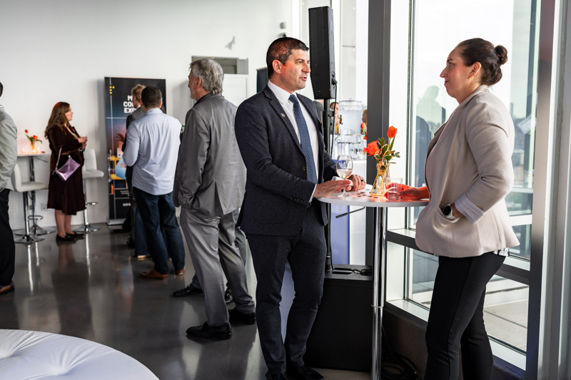 Business networking event: man in a suit chats with a woman at a high table by floor-to-ceiling windows, drinks and flowers on the table.