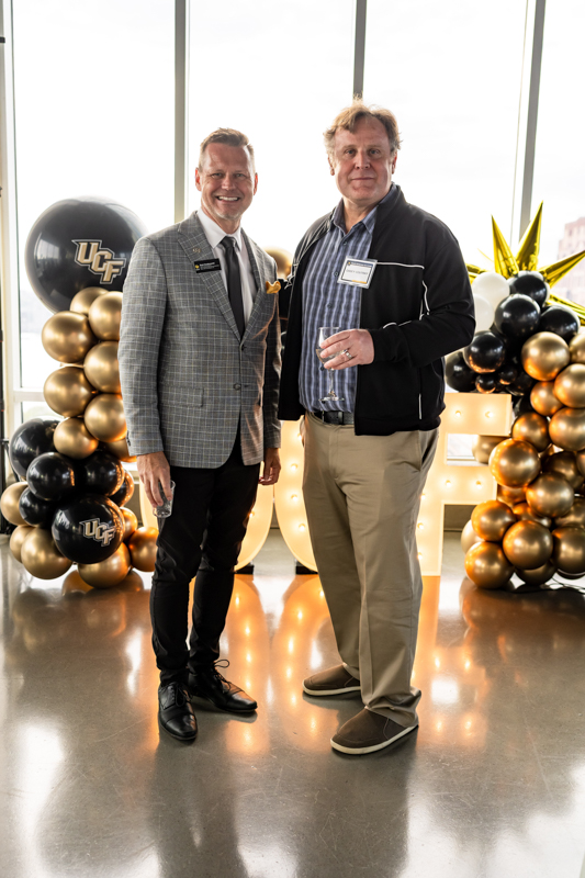 Two men pose for a photo at an event with black and gold UCF balloons and illuminated letters in a bright room, celebrating.