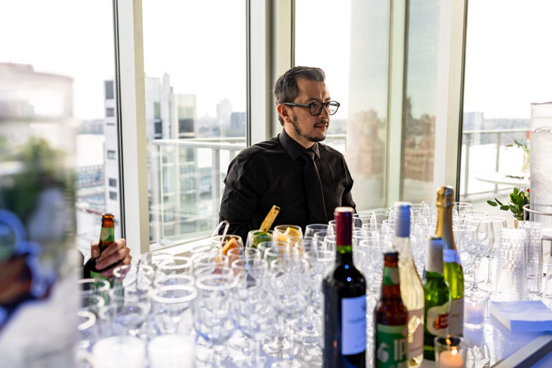 Man in a black shirt and tie sits at a reception table filled with wine glasses and bottles, with a city skyline visible through large windows behind him.