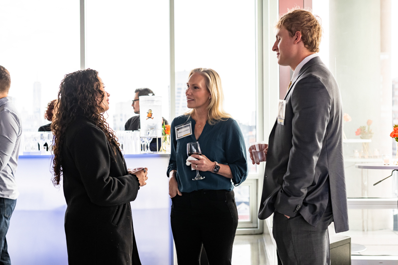 Three professionals in business attire chat and hold drinks near a bright event bar, name badges visible on their jackets/shirts.