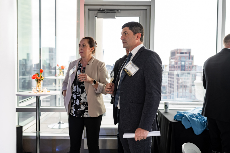 Two professionals in business attire stand near a glass door at a networking event, holding wine glasses with a city skyline in the background.