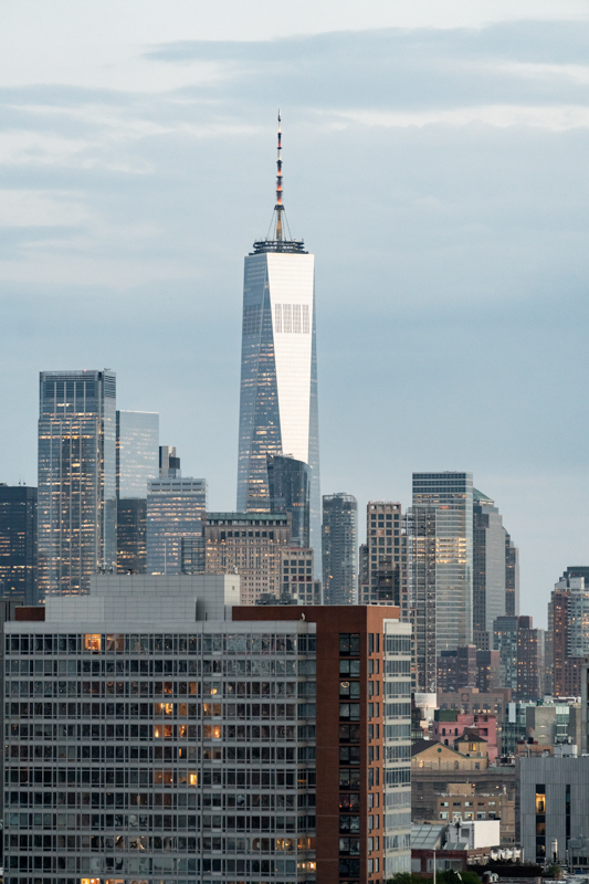 Skyline of Manhattan with the tall One World Trade Center rising above shorter buildings at dusk/light clouds overhead