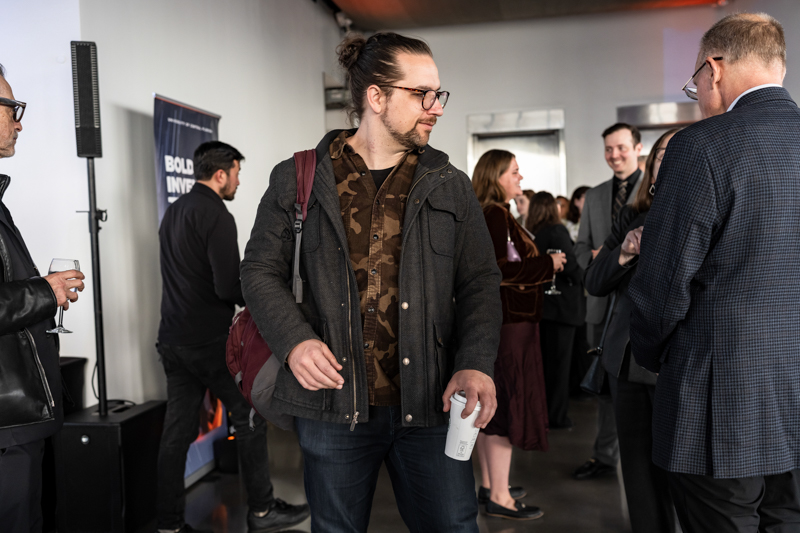 Man with a bun and glasses walking through a reception, holding a white cup; several people mingle in a modern event space in the background.