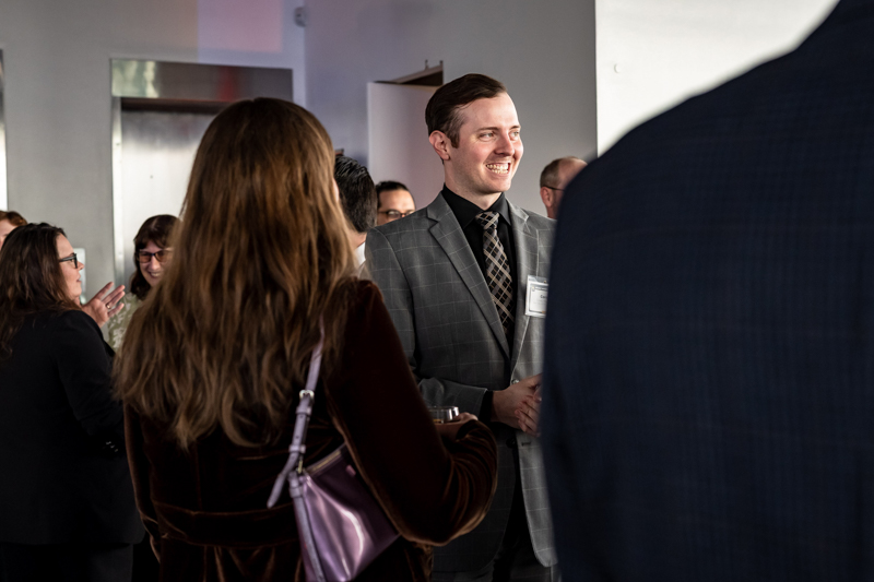 Smiling man in a gray plaid suit chats at a networking event, with other attendees in the background in conversation.