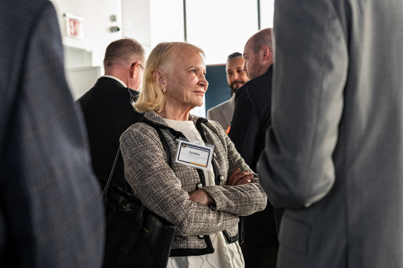 Senior woman with a name badge at a networking event, arms crossed, listening to conversations among attendees in a conference setting.