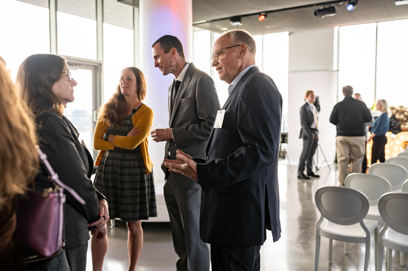 Professionals in business attire converse at a networking event in a bright, modern venue with empty chairs nearby.