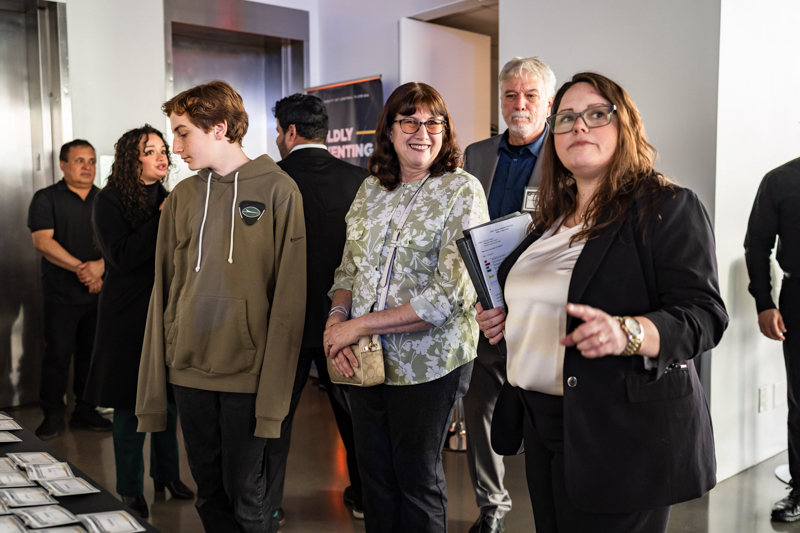 Group of adults at a reception, looking at a table of materials; a woman in a blazer gestures toward the table while others smile and chat.