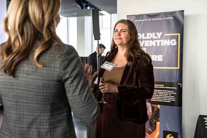 Woman with a name badge and champagne flute talks with another attendee at a conference, with a 'Boldly Inventing' banner in the background.