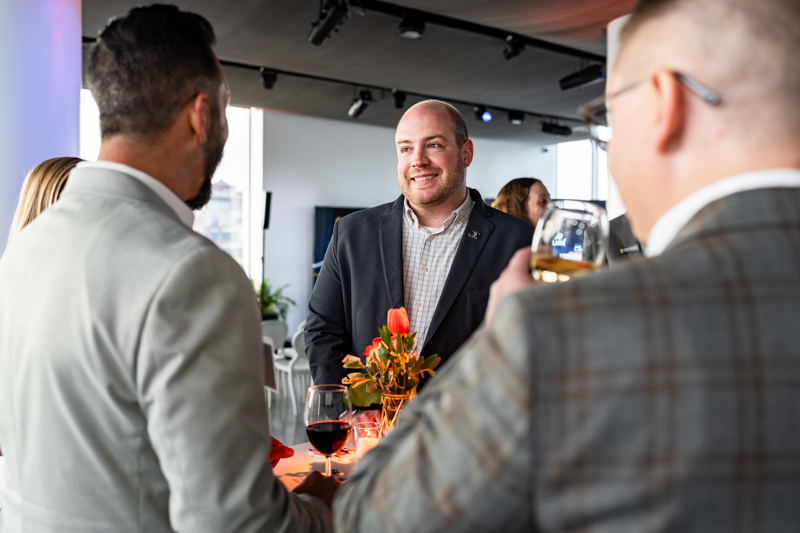 Professionals at a networking event chatting around a high-top table with wine glasses and an orange floral centerpiece.