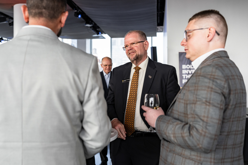 Businessmen in suits converse at a corporate networking event; central speaker wears an orange patterned tie as colleagues listen.