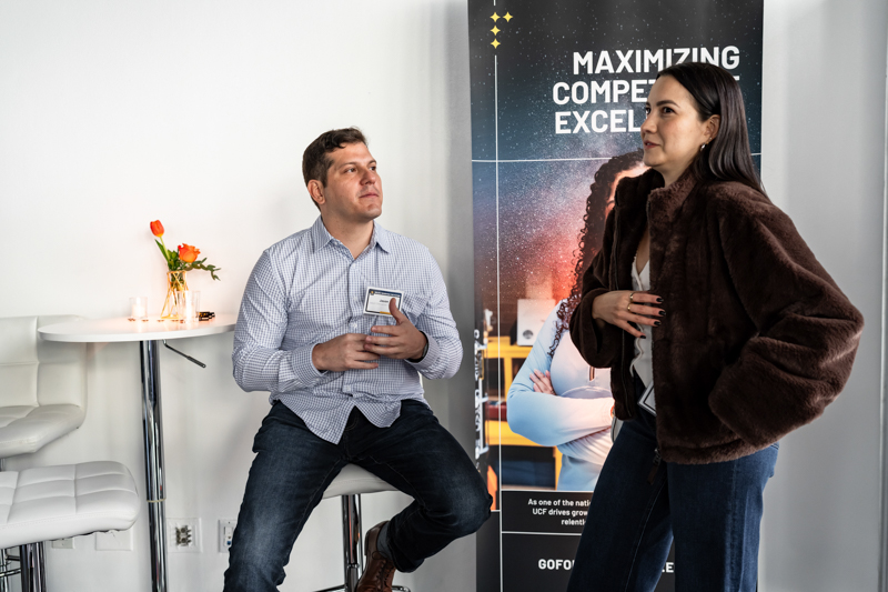 Man seated with a name badge talking to a woman in a brown fur coat beside a tall event banner with text behind them.