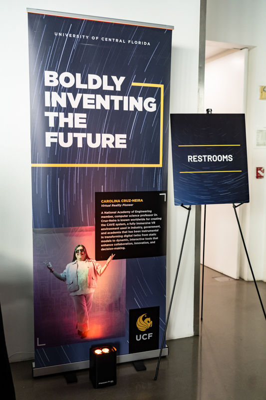 Tall banner reading 'Boldly Inventing the Future' for UCF, with a person pictured near the bottom and a separate 'Restrooms' sign beside it.