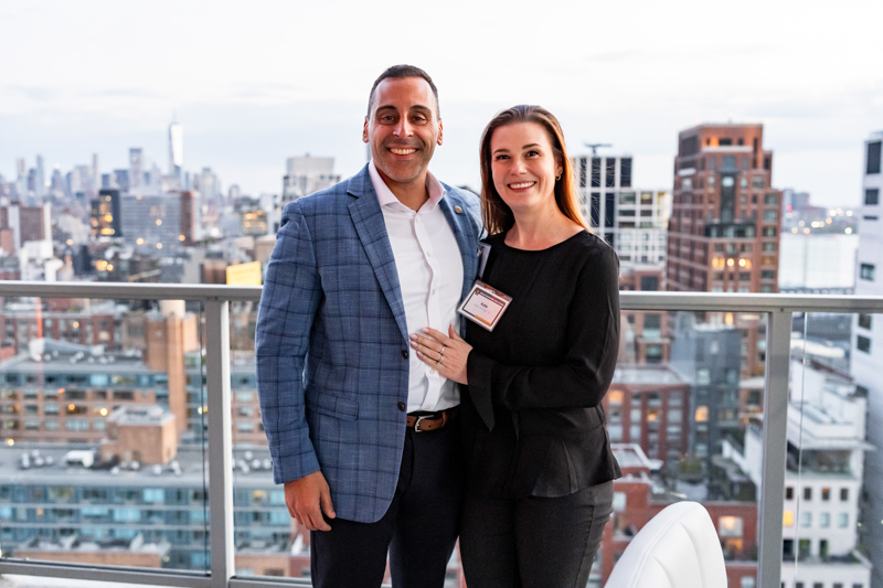 Smiling man in a blue blazer and woman with a name badge posing on a rooftop balcony with the city skyline behind them.