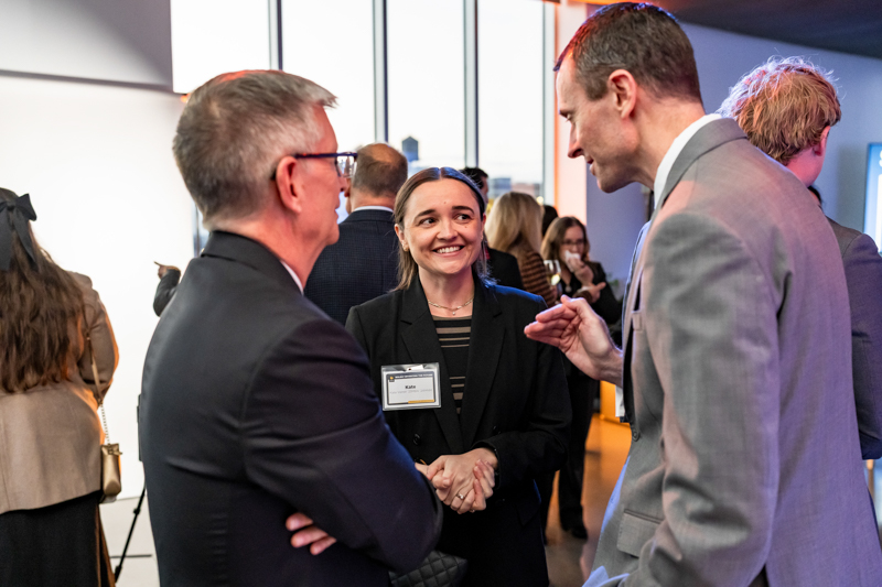 Two professionals shake hands and chat at a networking event, with a smiling woman wearing a name badge at the center.