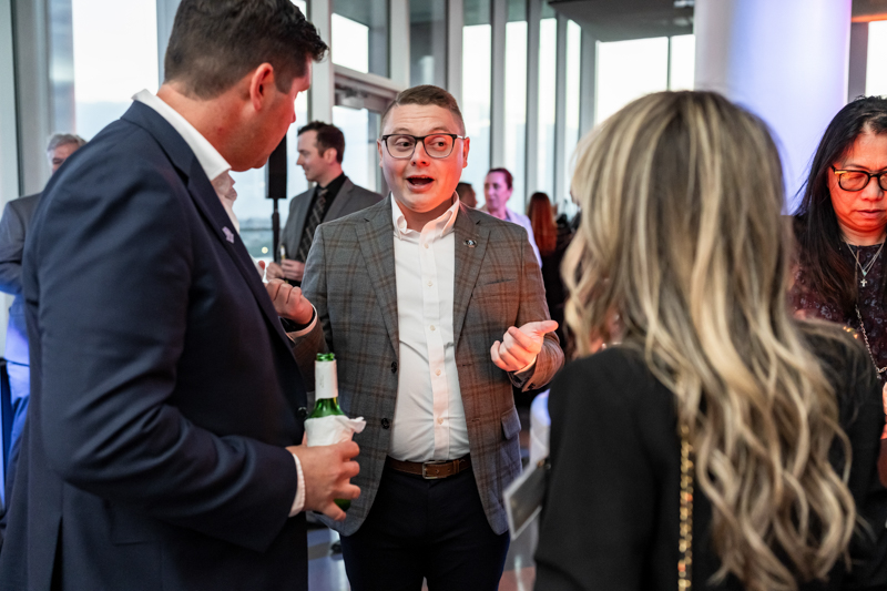 Man in a gray checkered blazer speaks with animated hand gestures at a wine reception.
