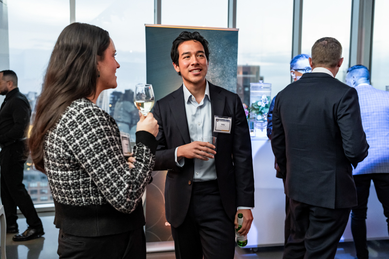 Networking at a corporate event: man in a suit with a name badge holds a drink and chats with a woman holding a wine glass; city skyline in the background.