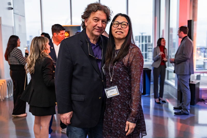 Two adults posing together at a conference lobby, smiling; the woman wears glasses and a name badge, the man in a dark blazer.