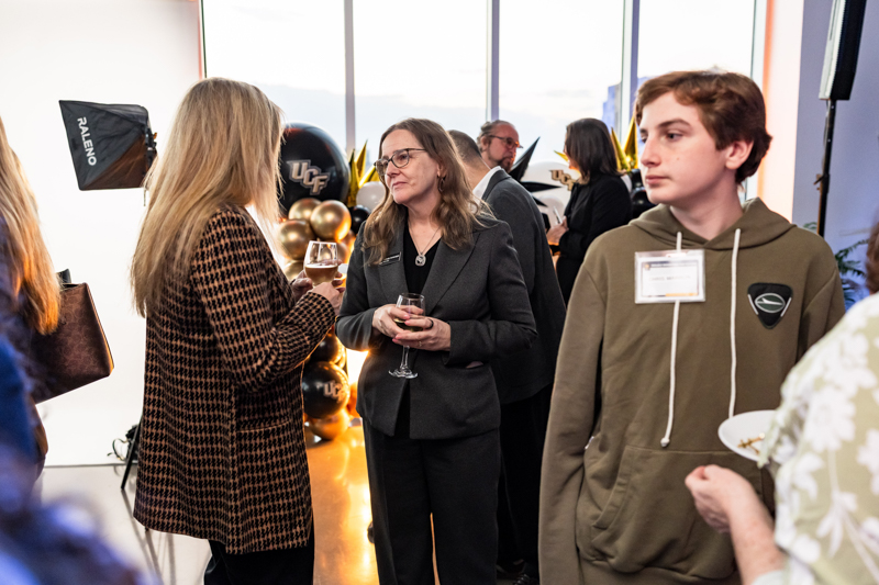 Two women chat at a reception, holding wine glasses, with gold balloons and a bright windowed backdrop behind them in a gallery-like space.