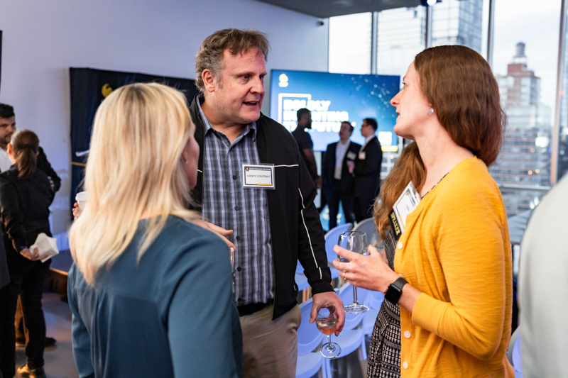 Professionals networking at a conference, two women and a man conversing with name badges in a brightly lit event space.