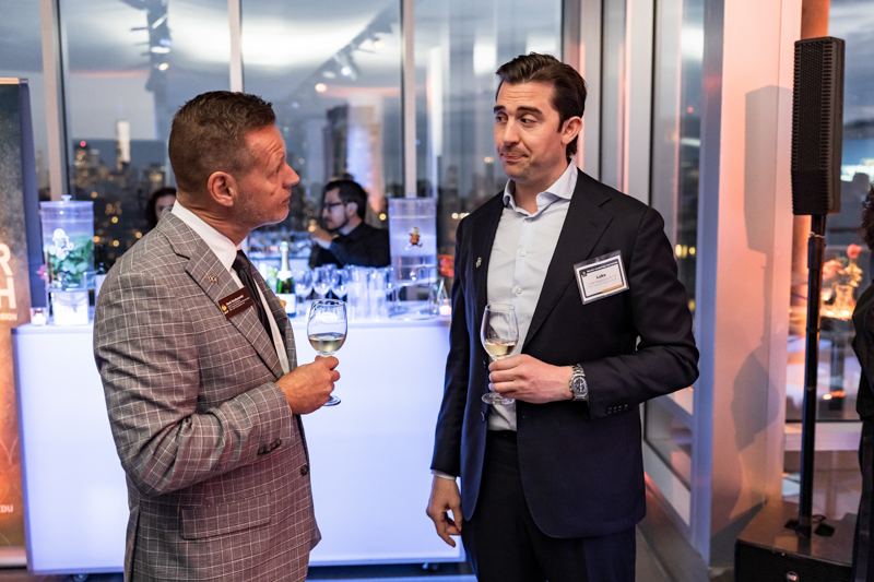 Two men in suits converse at a wine-tasting event, each holding a glass of white wine, in a modern venue with city lights outside.