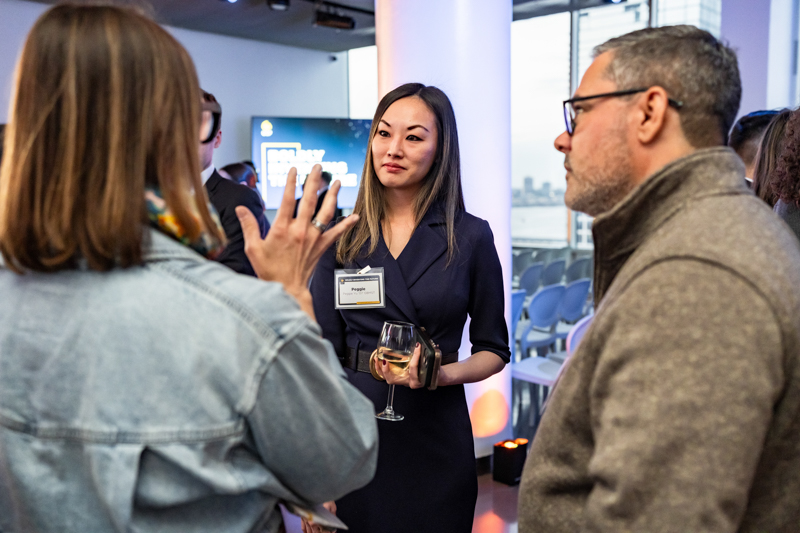 Professional woman in a navy dress holds a wine glass and listens during a networking event, wearing a name badge.
