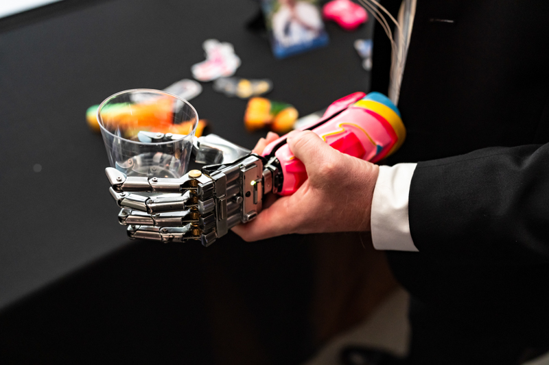 Close-up of a metallic prosthetic hand gripping a clear plastic cup, with a bright colorful toy in the background.