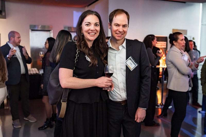A smiling woman in a black dress and a man in a suit pose together at a cocktail reception, holding a glass of wine and smiling at the camera.