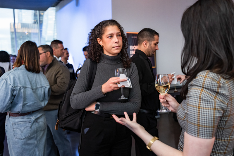 Woman in a gray turtleneck holds a wine glass and napkin while listening at a reception.