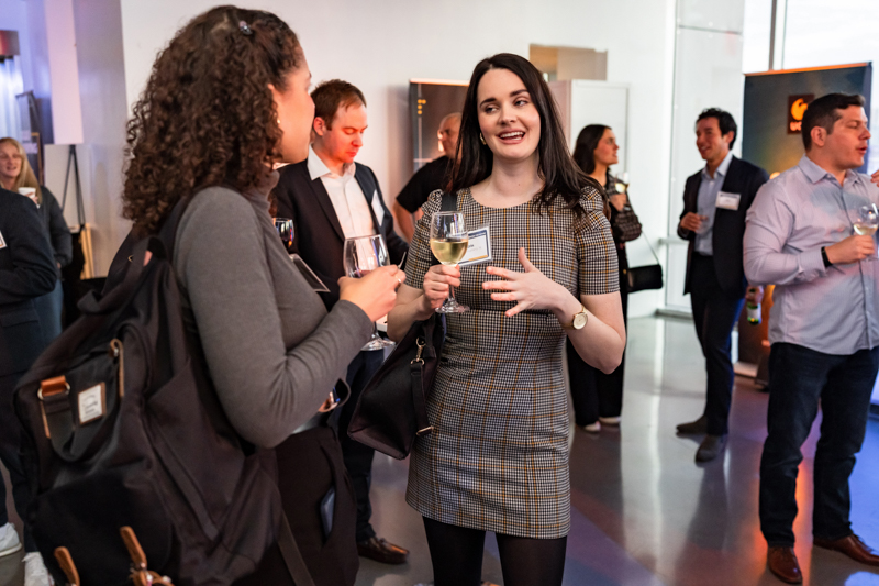 Two women converse at a networking event, holding wine glasses in a modern venue with other attendees in the background.
