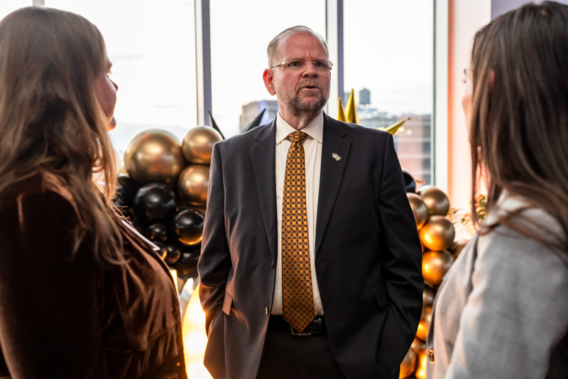 A man in a suit and orange tie speaks to two people at an event with gold and black balloons in the background.