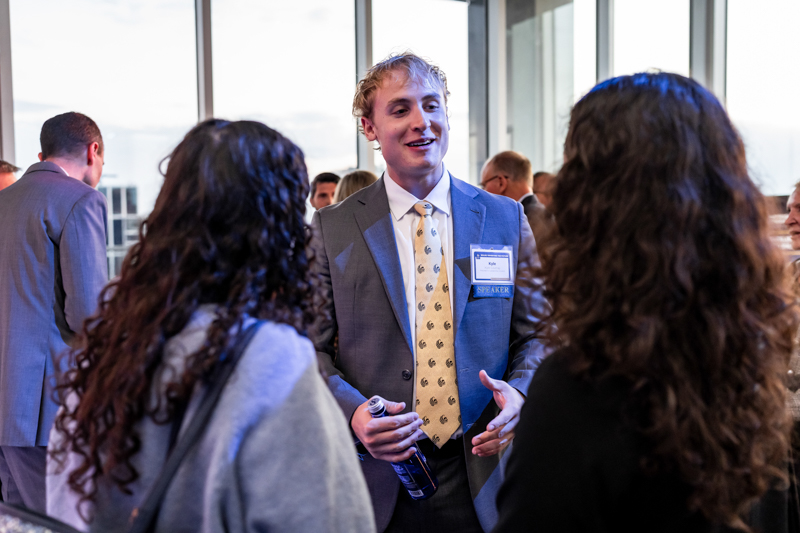 Young man in a gray suit and yellow tie talks with two attendees at a conference, wearing a speaker badge indoors by large windows.