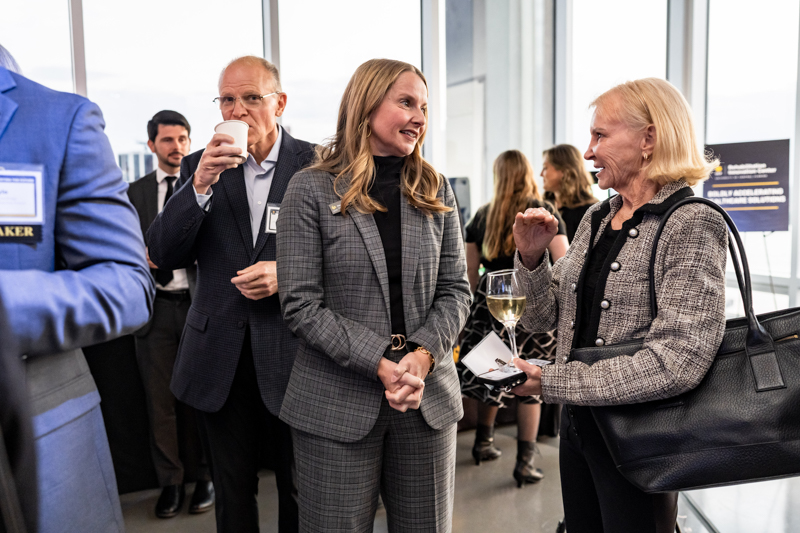 Professional reception scene: two women in business attire converse by large windows; one holds a wine glass while others chat nearby.