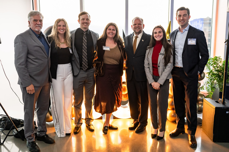 Eight professionals in business attire pose for a group photo at a modern office event, gold balloons in the background.