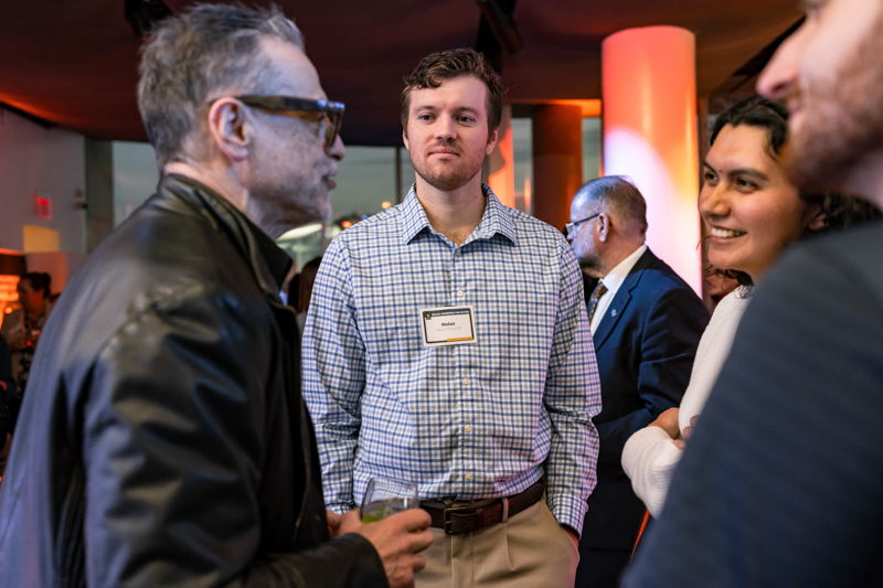Group of professionals chatting at a networking event, name badges visible, warm venue lighting in the background.