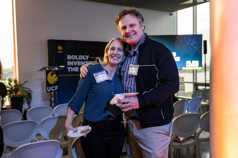 Two smiling adults pose together at an event, holding cocktails while wearing name badges, with a banner and chairs in the background.