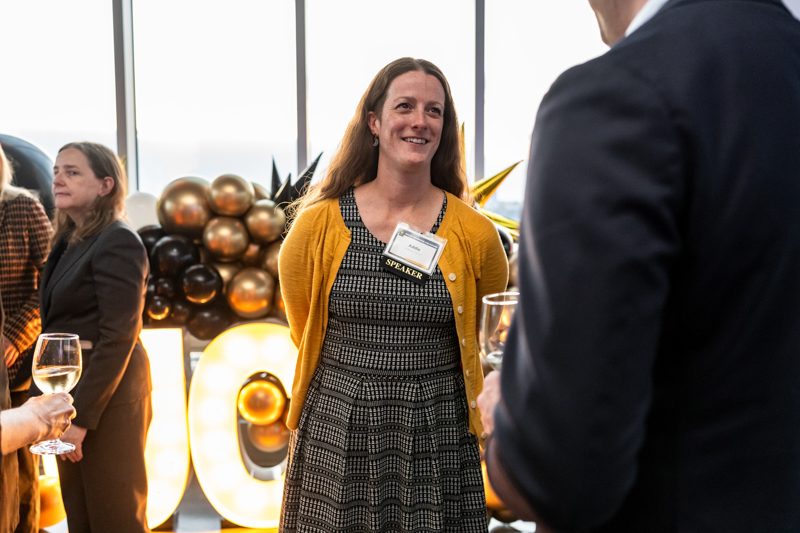 Smiling woman in a yellow cardigan wearing a speaker badge chats with a man at a reception, with balloons and warm lights in the background.