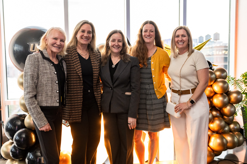 Five women in business attire posing together indoors, with black and gold balloons in the background.