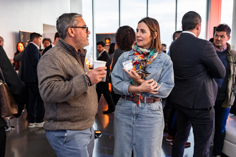 Two adults converse at a casual networking event, the man holding a coffee cup while the woman in a denim dress listens, scarf wrapped around her neck, badge in hand.