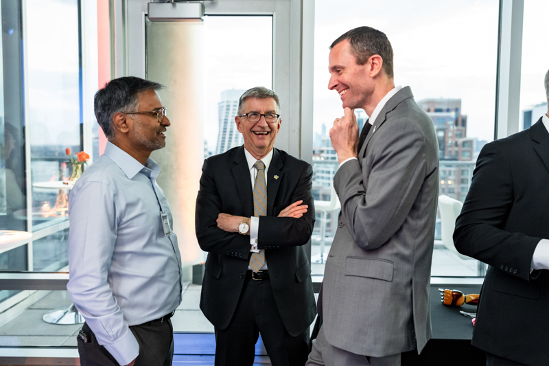 Three businessmen in suits chat and smile by a window overlooking a city skyline in a modern office setting.