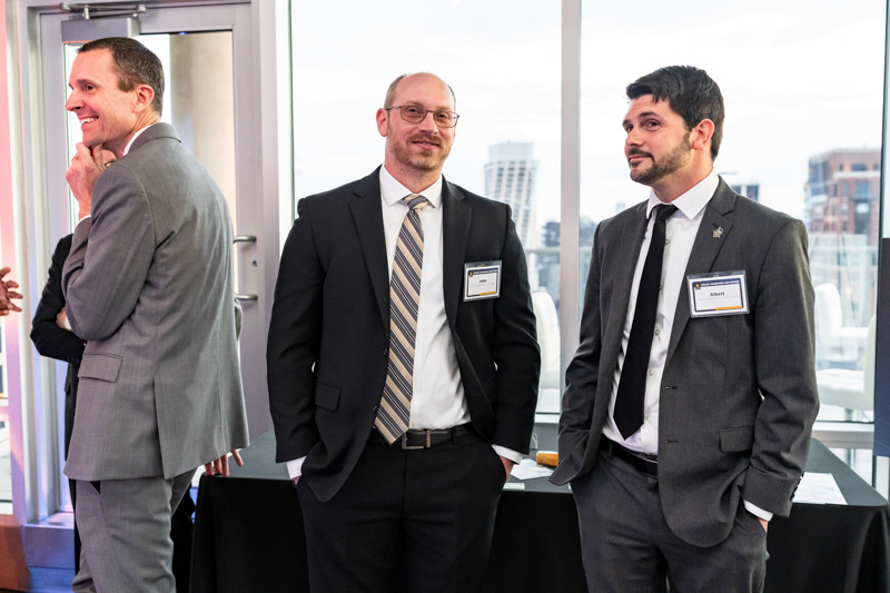 Three businessmen in suits with name badges chat at a conference by large windows.