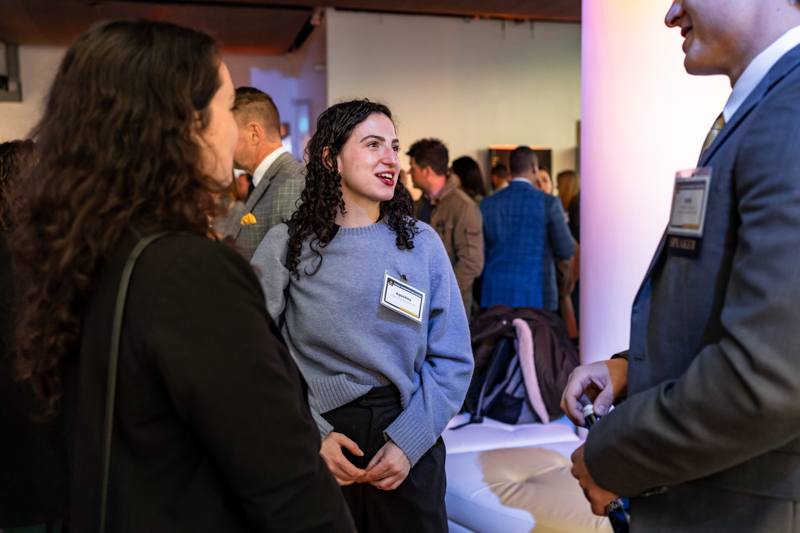 Woman in a blue sweater talks with colleagues at a networking event; name tag visible on her chest.