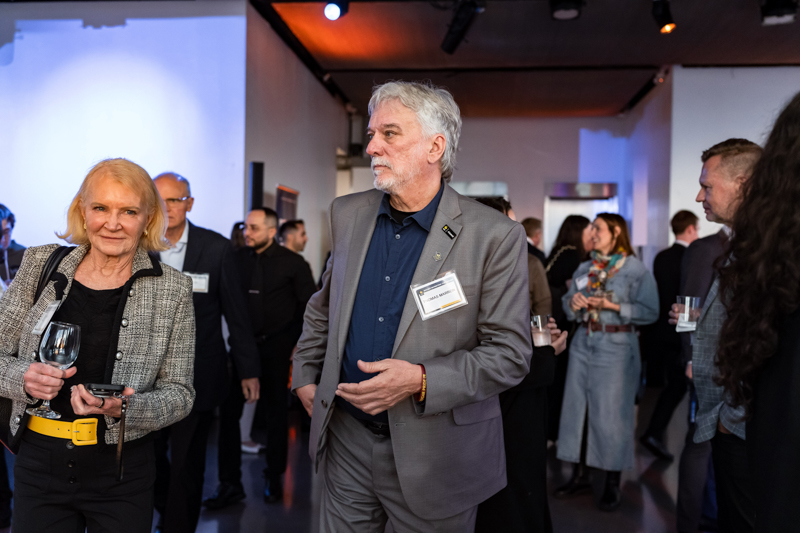 Networking event image: attendees in business attire mingle, a man in a gray suit with a name badge in the foreground, others holding drinks.