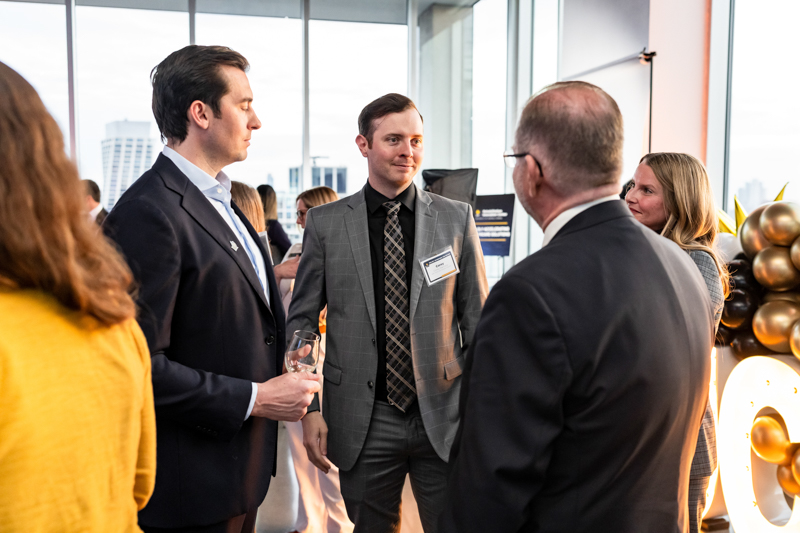 Professionals in suits chat at a networking event in a bright office, with balloons in the background and attendees wearing name badges.
