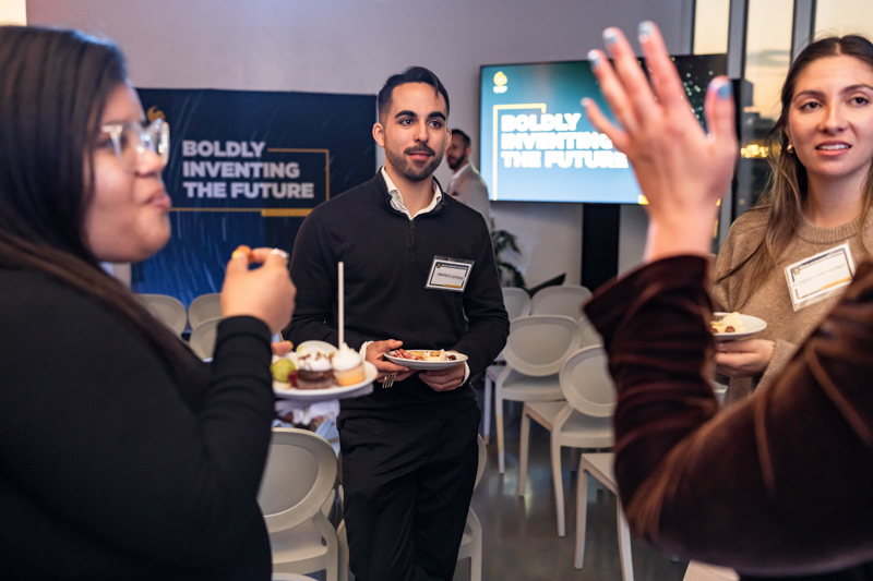Young professionals at a conference reception, plates of dessert in hand, chatting near a banner reading 'Boldly Inventing the Future.'