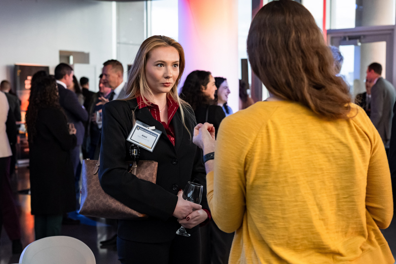 Businesswoman in a black suit with a name badge talks to a colleague at a networking event, holding a wine glass and clutch.