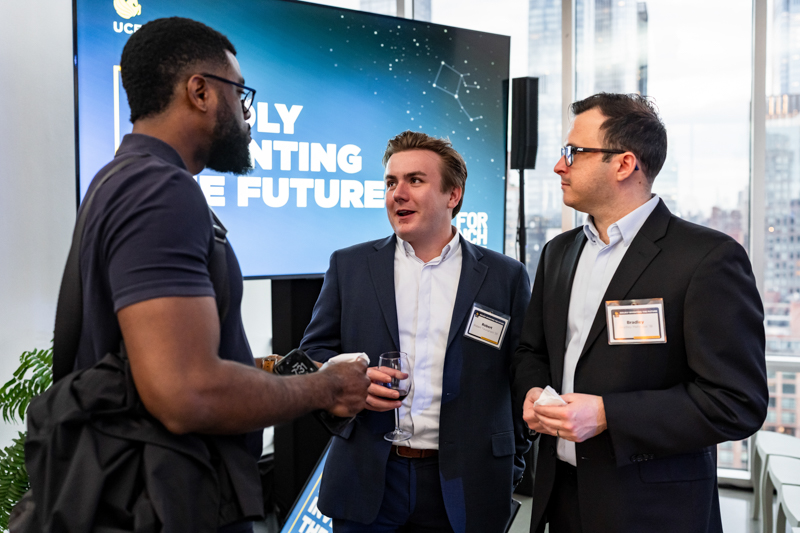 Three men in business attire converse at a conference; one man on the left with a backpack talks to two men in suits, one holding a wine glass.