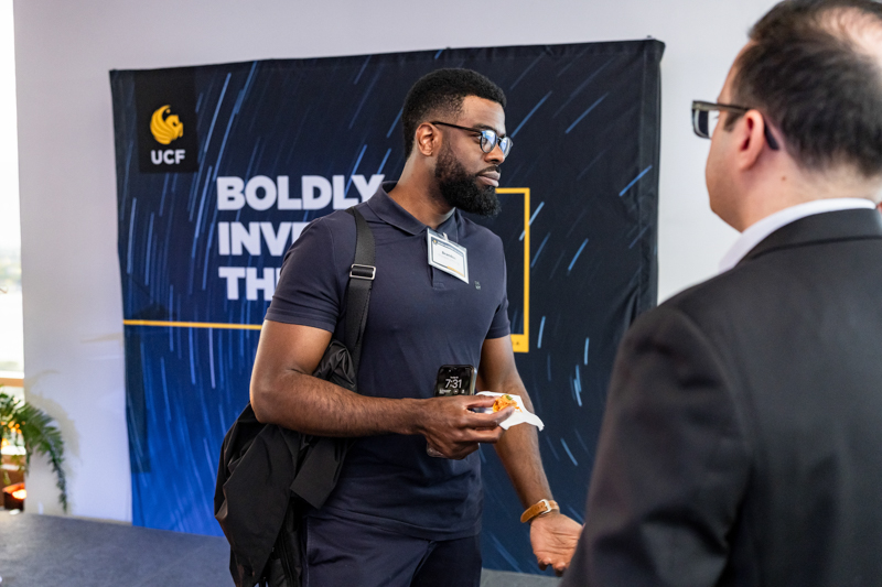 Conference attendee with beard in navy polo hands food to a man in a suit, backdrop shows 'Boldly Involv...'; badge visible on chest.